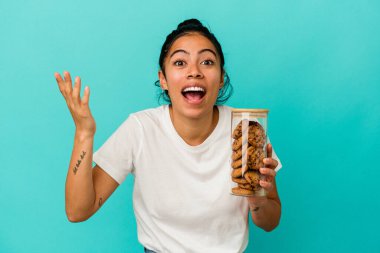 Mavi arka planda izole edilmiş kurabiye kavanozunu tutan genç Latin kadın hoş bir sürpriz alıyor, heyecanlı ve el kaldırıyor..