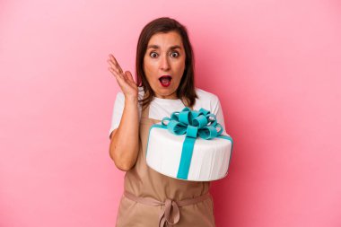 Orta yaşlı pasta şefi kadın elinde bir pasta tutuyor mavi arka planda izole edilmiş şaşırmış ve şok olmuş..