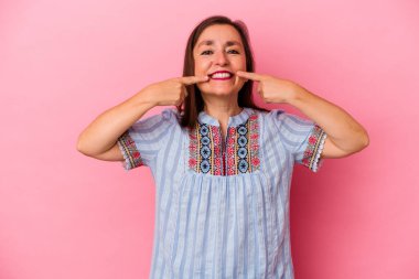 Orta yaşlı beyaz kadın pembe arka planda izole edilmiş gülücükler içinde, parmaklarını ağıza doğrultuyor..