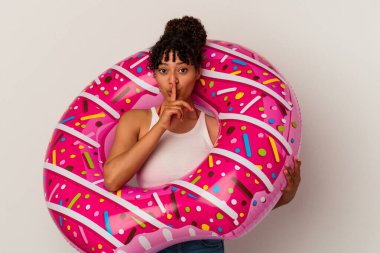 Beyaz arka planda izole edilmiş şişme bir donut tutan genç melez kadın ya sır saklıyor ya da sessizlik istiyor..