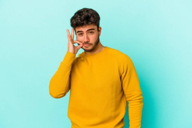 Genç beyaz adam mavi arka planda izole edilmiş ve parmakları dudaklarında bir sır saklıyor..