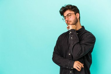 Mavi arka planda izole edilmiş genç beyaz adam şüpheli ve şüpheci bir ifadeyle yana bakıyor..
