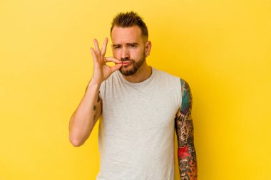 Sarı arka planda izole edilmiş genç dövmeli beyaz adam dudaklarında parmaklarıyla bir sır saklıyor..