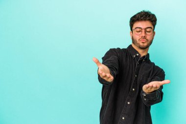 Kafkasyalı genç adam mavi arka planda izole edilmiş dudaklar kıvrılıyor ve hava öpücüğü göndermek için avuç içleri tutuyor..