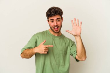 Beyaz arka planda izole edilmiş genç beyaz bir adam gülümseyerek beş numarayı parmaklarıyla gösteriyor..