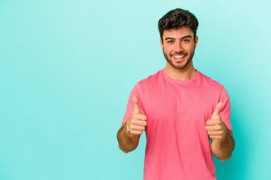 Mavi arka planda izole edilmiş genç beyaz adam gülümsüyor ve başparmağını kaldırıyor.