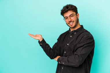 Mavi arka planda izole edilmiş genç beyaz bir adam bir palmiye üzerinde fotokopi alanı ve belinde bir el daha gösteriyor..