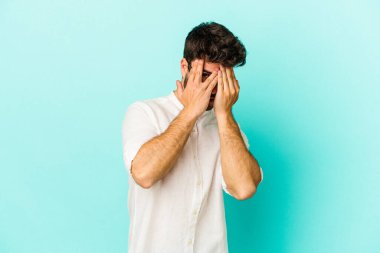 Kafkasyalı genç adam mavi arka planda izole edilmiş. Korkmuş ve gergin parmaklarıyla göz kırp..