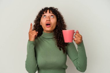 Genç melez bir kadın beyaz arka planda izole edilmiş bir kupa tutuyor ve ağzı açık bir şekilde üst tarafı işaret ediyor..