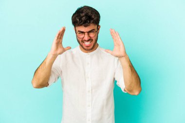 Mavi arka planda izole edilmiş genç beyaz adam öfkeyle bağırıyor..