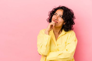 Genç kıvırcık Latin kadın pembe arka planda izole edilmiş rahat bir şekilde fotokopi odasına bakan bir şeyi düşünüyor..