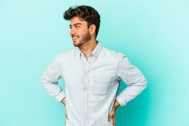 Kafkasyalı genç, mavi arka planda izole edilmiş. Kendine güvenen, kalçaları tutan biri..