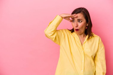 Orta yaşlı, beyaz bir kadın pembe arka planda izole edilmiş, uzaklara bakıyor ve elini alnına koyuyor..