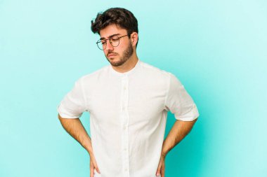Kafkasyalı genç adam mavi arka planda izole, kafası karışmış, şüpheli ve güvensiz hissediyor..
