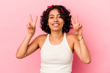 Pembe arka planda izole edilmiş genç kıvırcık Latin kadın zafer işareti gösteriyor ve gülümsüyor..