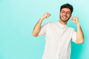 Mavi arka planda izole edilmiş genç beyaz bir adam zaferden sonra yumruğunu kaldırıyor, kazanan konsept..