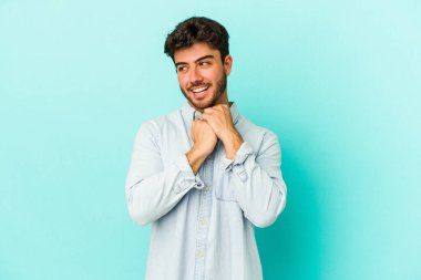 Mavi arka planda izole edilmiş genç beyaz adam ellerini çenesinin altında tutuyor, mutlu bir şekilde yana bakıyor..