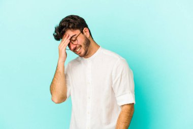 Mavi arka planda izole edilmiş genç beyaz adam kameraya parmaklarıyla göz kırpıyor, yüzünü kapatmaktan utanıyor..