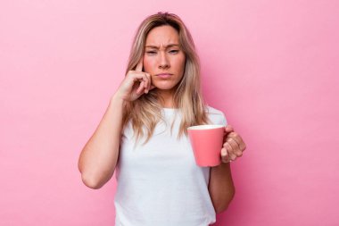 Genç Avustralyalı bir kadın pembe arka planda izole edilmiş pembe bir kupa tutuyor. Parmağıyla tapınağı işaret ediyor, düşünüyor, bir göreve odaklanıyor..
