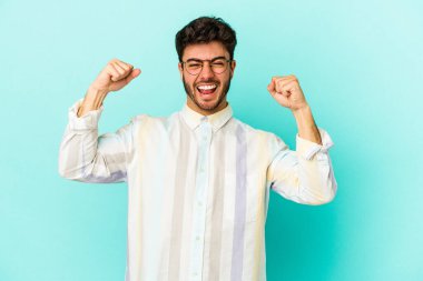 Kafkasyalı genç adam mavi arka planda izole edilmiş kaygısız ve heyecanlı tezahüratlar yapıyor. Zafer konsepti.