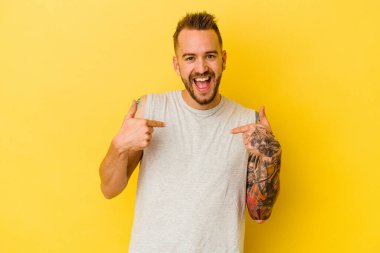 Sarı arka planda izole edilmiş genç dövmeli beyaz adam parmağını gösterip gülümseyerek şaşırdı..