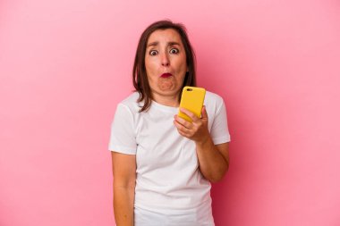 Elinde cep telefonu olan orta yaşlı beyaz bir kadın pembe arka planda izole edilmiş omuzlarını silkeliyor ve gözleri açık..