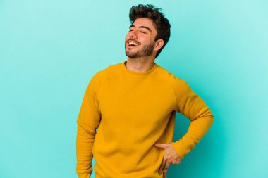 Kafkasyalı genç adam mavi arka planda izole edilmiş rahat ve mutlu bir şekilde gülüyor, boynunda dişlerini gösteriyor..