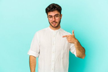 Mavi arka planda izole edilmiş genç beyaz adam bir gömlek fotokopi alanını işaret ediyor, gururlu ve kendinden emin.