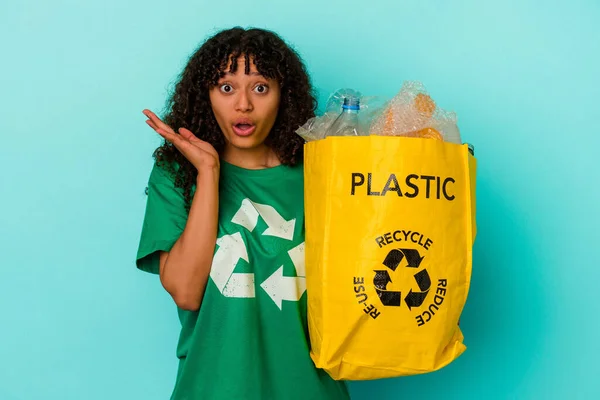 年轻的混血女子拿着一个蓝色背景的回收塑料袋 感到惊讶和震惊 — 图库照片