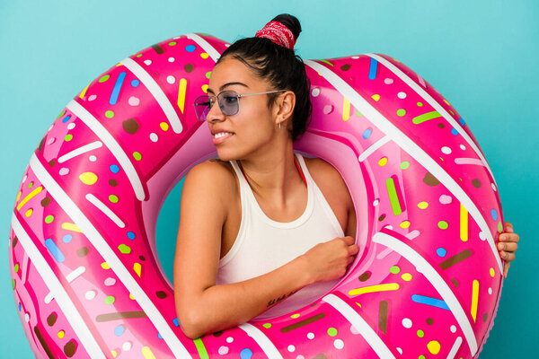 Young latin woman holding an inflatable donut isolated on blue background looks aside smiling, cheerful and pleasant.