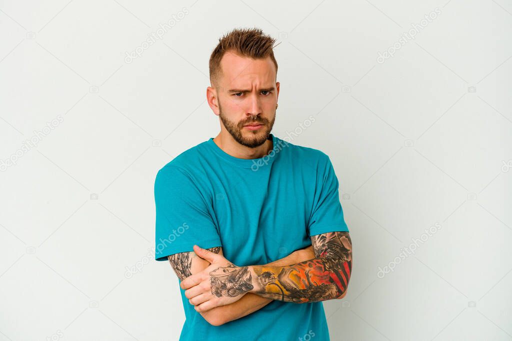 Young tattooed caucasian man isolated on white background frowning face in displeasure, keeps arms folded.
