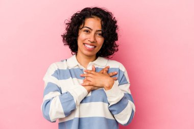 Pembe arka planda soyutlanmış genç kıvırcık Latin kadın gülüyor, ellerini kalbe koyuyor, mutluluk kavramını koruyor..
