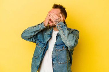 Sarı arka planda izole edilmiş genç dövmeli beyaz adam elleriyle gözlerini kapatmaktan korkuyor..
