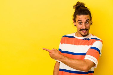 Sarı arka planda izole edilmiş uzun saçlı beyaz bir adam gülümsüyor ve boş alanda bir şeyler gösteriyor..