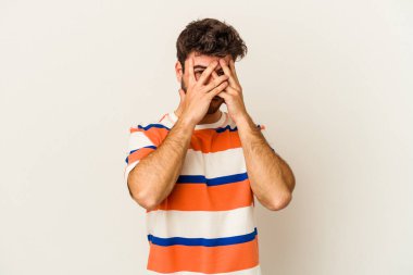 Beyaz arka planda izole edilmiş genç beyaz bir adam parmaklarının arasından korkmuş ve gergin bir şekilde göz kırpıyor..