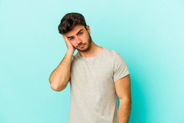 Kafkasyalı genç adam mavi arka planda izole edilmiş. Yorgun ve uykulu. Elini başının üstünde tutuyor..