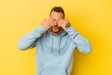 Sarı arka planda izole edilmiş genç dövmeli beyaz adam elleriyle gözlerini kapatmaktan korkuyor..