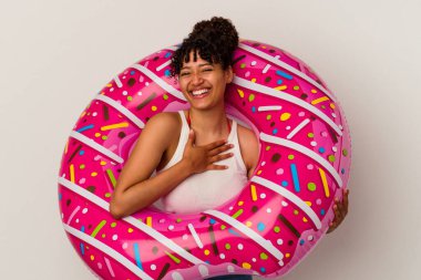 Beyaz arka planda izole edilmiş şişme bir donut tutan genç bir ırk kadını kahkahalarla gülüyor ve elini göğsüne koyuyor..