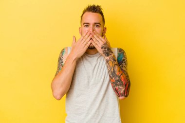 Sarı arka planda izole edilmiş genç dövmeli beyaz adam şok olmuş, ağzı ellerle kaplanmış, yeni bir şey keşfetmeye hevesli..