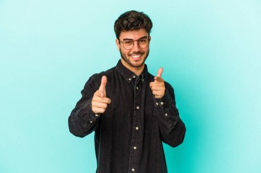 Mavi arka planda izole edilmiş genç beyaz adam parmaklarıyla ön tarafı işaret ediyor..