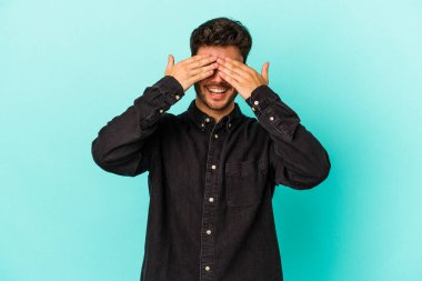 Mavi arka planda soyutlanmış genç beyaz bir adam elleriyle gözlerini kapatıyor, geniş bir gülümsemeyle bir sürpriz bekliyor..