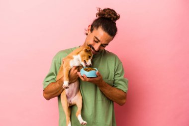 Genç beyaz adam köpeğini tutuyor ve yemeği pembe arka planda izole edilmiş.