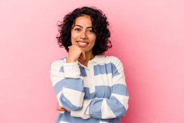 Genç kıvırcık Latin kadın pembe arka planda izole edilmiş mutlu ve kendinden emin gülümsüyor, elleriyle çenesine dokunuyor..