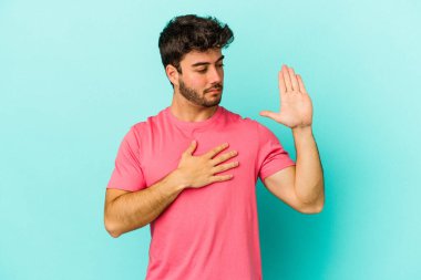 Kafkasyalı genç adam mavi arka planda izole edilmiş yemin ediyor, elini göğsüne koyuyor..