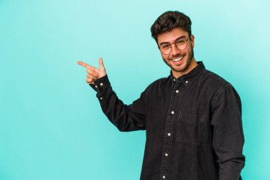 Mavi arka planda izole edilmiş genç beyaz adam işaret parmağıyla işaret ederek gülümsüyor..