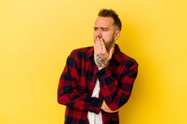 Sarı arka planda izole edilmiş genç dövmeli beyaz adam esniyor. Ağzı eliyle kapatan yorgun bir hareket sergiliyor..