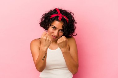 Pembe arka planda izole edilmiş genç kıvırcık Latin kadın yumruğunu kameraya gösteriyor, agresif yüz ifadesi..