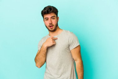 Mavi arka planda izole edilmiş genç beyaz adam parmağını göstererek şaşırdı, geniş bir gülümsemeyle..