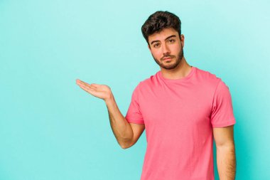 Mavi arka planda izole edilmiş genç beyaz bir adam bir palmiye üzerinde fotokopi alanı ve belinde bir el daha gösteriyor..