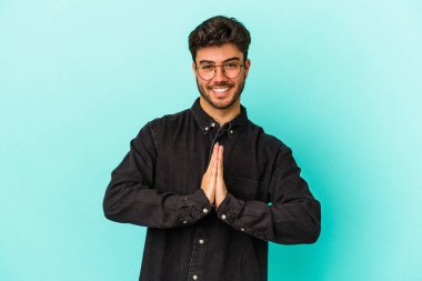 Mavi arka planda izole edilmiş genç beyaz bir adam el ele tutuşup, ağzının kenarında dua ediyor, kendinden emin..